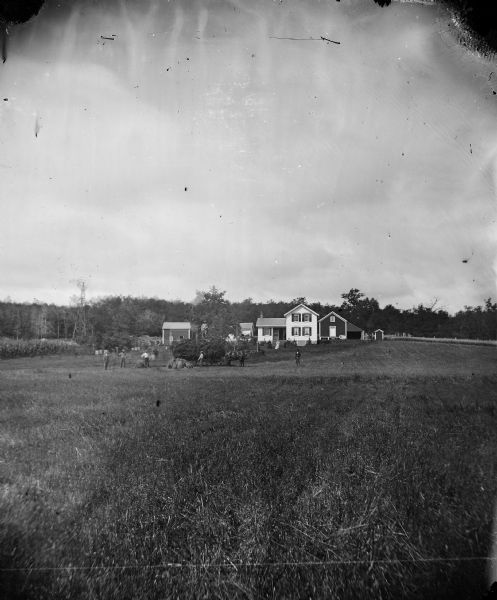 Men in a grain field are piling hay or straw onto a wagon as one man stands on top of the pile. On the left is a windmill on a wooden tower and behind are several small buildings, including a granary and a privy. A woman stands in front of an upright and wing frame house. A cornfield is on the left and possibly an adjoining beehive.