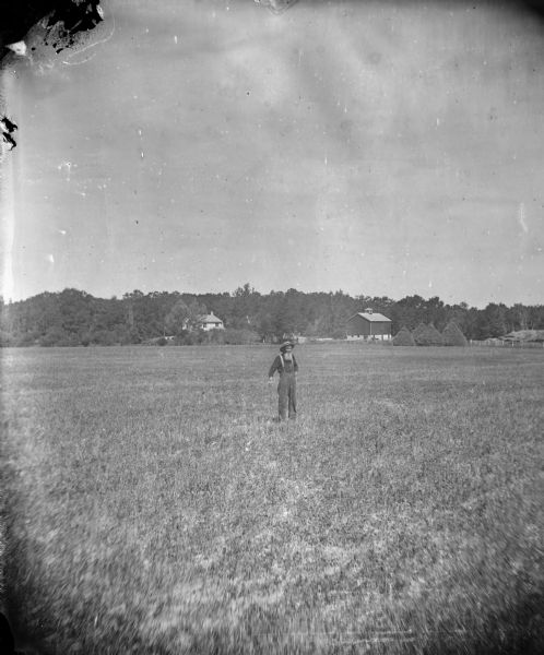 A bearded man stands with a cane in a field. Behind him are large haystacks, a barn with a cupola and a house in front of a forested area.