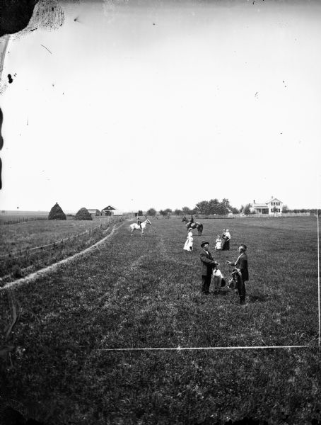Two men stand in the foreground on the verge of shaking hands. Behind them are women on horseback with haystacks and a farmstead in background.