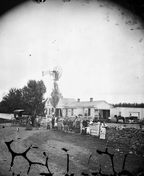 A large group of men, women and children stand in front of an elaborate wood and stone fence. A horse and carriage stand at the left and behind them is a windmill with two men posing on top. On the right are other carriages and wagons and what appears to be a commercial building attached to a house with screen doors. There may be a wagon works or other business here.