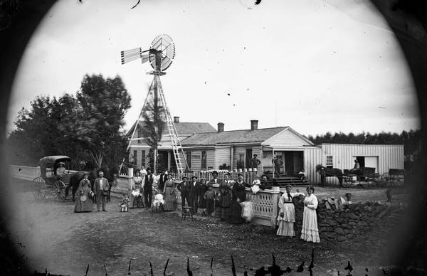 A large group of men, women and children stands in front of an elaborate wood and stone fence. A horse and carriage stand at the left and behind them a windmill with a man posing on top. On the right are other carriages and wagons and what appears to be a commercial building attached to a house with screen doors. There may be a wagon works or other business here.