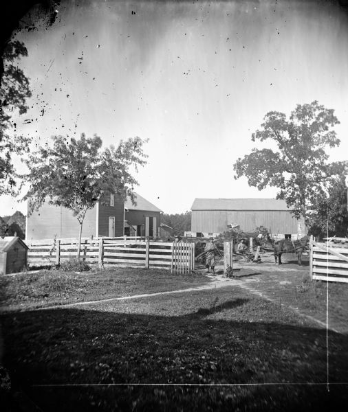 Men and horses stand in the fenced-in barnyard of the G. Larsen farm.  A mower and a self-raking reaper can also be seen.  In the background is a barn and another building.