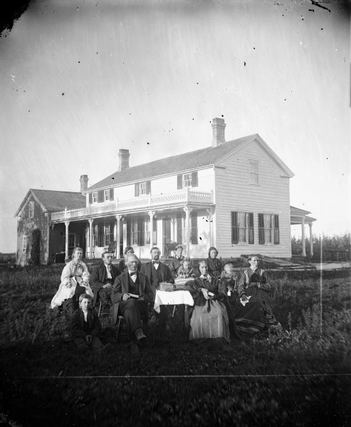 The G. Larsen Family sits in their front yard. Behind them a sewing machine can be seen on the porch of their two-story house.