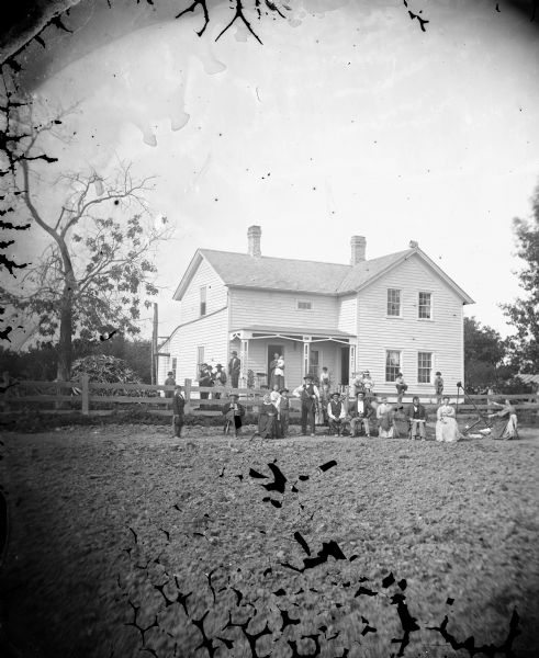 View across bare ground towards a large extended family of 26 people who are sitting and standing along a fence in the front yard of a wood frame house; Lars Davidson Reque. On the right a woman is operating a spinning wheel, and on the left is a large pile of wood scraps.