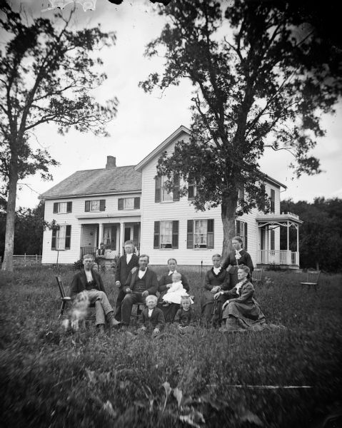 The Ole Farner family is posing in their yard. Two men, a dog and a woman with an infant are sitting on the left, with a boy standing behind them, and two children sitting in the grass in front of them. On the right one woman is standing behind two women sitting, with one of them holding a hand fan. Behind the group two men, one sitting, one obscured by the boy in front, are in front of the porch of a frame house which has small second-story windows on that wing. There is a porch entryway on the right side of the house. All the windows have shutters. There is one empty chair on the left near the group, and two empty chairs in the grass on the right.