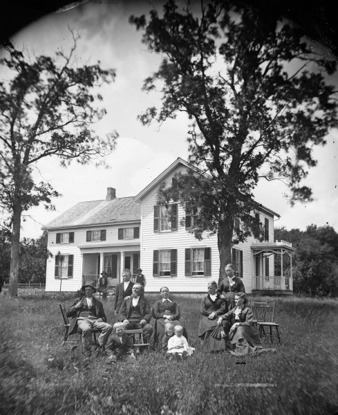 The Ole Farner family is posing in their yard. Two men and a woman are sitting on the left, with a boy standing behind them, and three children sitting in the grass in front of them. On the right one woman is standing behind two women sitting, with one of them holding an open hand fan. Behind the group two men, one sitting, one standing, are in front of the porch of a frame house which has small second-story windows on that wing. There is a porch entryway on the right side of the house. All the windows have shutters.