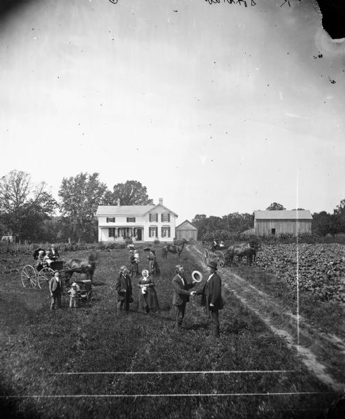 In the foreground, Erik Jensen, a minister with his hat in hand, shakes K. Doxstad's hand. Nearby a toddler pulls his own baby carriage. Two carriages, a boy on a horse and various people are arranged in the background with a farm house and outbuilding in the distance. Clinton is located in the Jefferson Prairie settled by Norwegian immigrants.