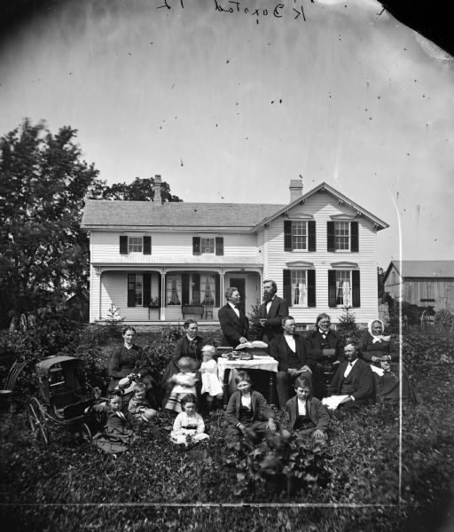 The K. Doxstad family is posed in their yard around a table covered with heavy books. A baby carriage is on the left and an older woman with her knitting in her lap on the right. Behind them is a frame house, a carriage and farm buildings. The taller man is the Reverend Erik Jensen. Clinton is located in the Jefferson Prairie settled by Norweigan immigrants.