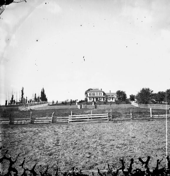 Long view of 10 members of a family in a large fenced yard. Behind them is a substantial two-story bracket style frame house with a widow's walk at the top of its hip roof.