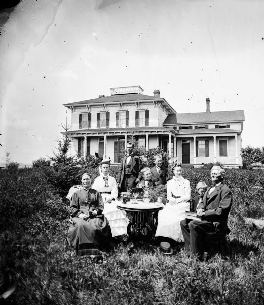 A family is seated around a table in their yard, with two of the women dressed in white. Behind them is a substantial two-story bracket style frame house with a widow's walk at the top of its hip roof.