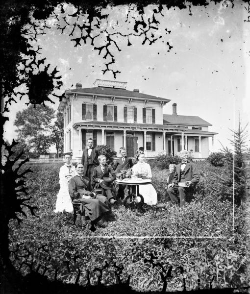 A family is posed in a yard around a table, with a boy holding his hat. Behind them is a substantial two-story bracket style frame house with a widow's walk at the top of its hip roof.
