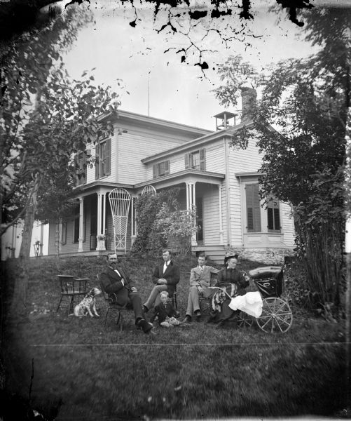 A husband and wife are posed with three sons, an infant in a baby carriage, and a Dalmatian dog, in front of a frame house with a stone foundation, shutters closed in a bay window on the side, a trellis in front and a large dinner bell on the roof.