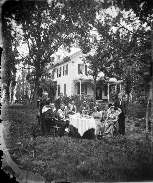 The Reverend Jakob Aall Ottesen (1825-1904), his wife Cathinka Doderlein Ottesen, their family and visitors are posing in a yard around a table set for coffee. On the left, Jakob Ottesen is holding a long-stemmed pipe, and behind him a ladder is leaning against a tree. The frame house behind them is surrounded by large trees and vines which have been trained to grow on a porch that has some carpenter's lace.