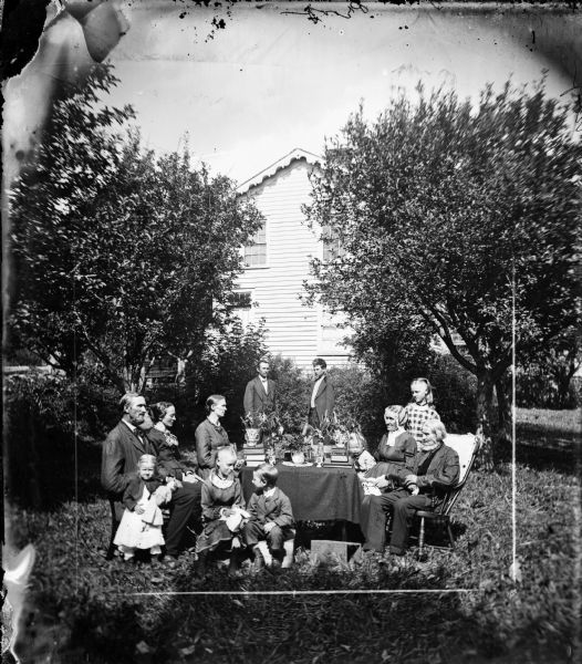 A family is gathered around a table loaded with books and potted plants. Children sit on soap boxes in front, including a girl with a doll. The frame house behind has nice roof trim.