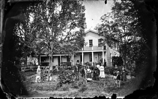A family is gathered in a yard playing croquet. Nearby a child sits on a rocking horse and holds onto a small wagon. Behind that are some women and another child with a baby buggy. The frame house has a railing on its porch roof and latticework in front. This is the Spring Prairie parsonage, home of Herman Amberg Preus, his wife Caroline Keyser Preus and their children.