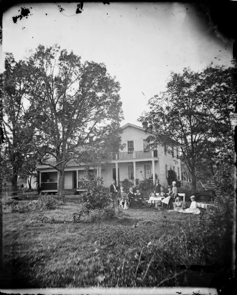A family is gathered in a yard around a table with a child on a rocking horse. The frame house behind them has a railing on its porch roof and latticework. A rustic wood bench sits under a tree on the right. This is the Spring Prairie parsonage, home of Herman Amberg Preus, his wife Caroline Keyser Preus and their many children.