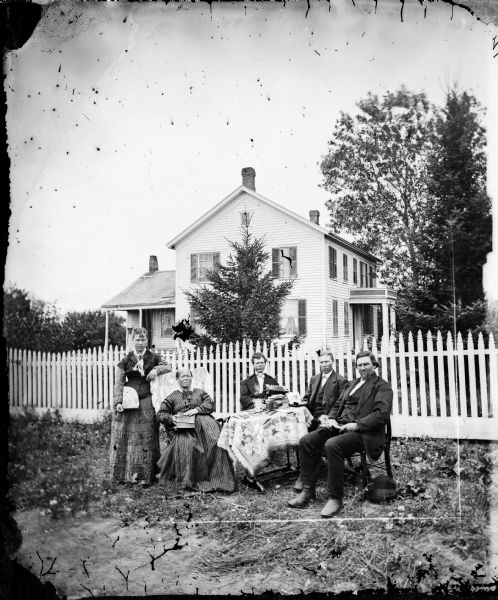 Three men and two women sit around a table in a yard. Behind them is a picket fence and a wood frame house. One woman holds a fan, another a book, while the man fingers a concertina.
