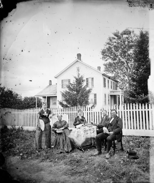 Three men and two women sit around a table in a yard. Behind them is a picket fence and a wood frame house. One woman holds a fan, another a book. One man fingers a concertina and another holds a newspaper.