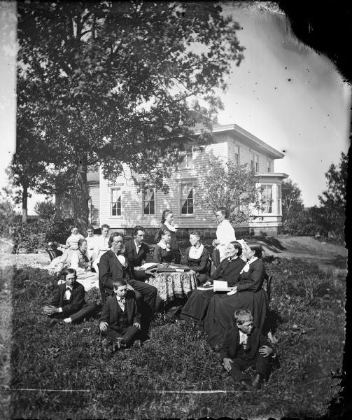 The Neils Hallanger family is posed in their yard around a table piled with books. One lady is posed with a parasol and servant women sit on a sofa in the left rear. The frame house has a bay window and a stone foundation behind. The people are carefully posed and face right or left, but no one looks directly at the camera. Gjertrud Amundsen Dahl, the niece of Neils and Brita Hallanger and wife of A.L. Dahl stands in the right rear.