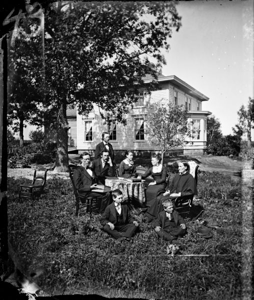 The Neils Hallanger family is gathered in their yard around a table with books on it and a love seat behind. The frame house has a bay window and a stone foundation. From the left, the people are Neils T. Hallanger, his sons Thomas and John (standing), his daughter Magdalena, daughter-in-law Sophia Foss Hallanger and his wife Brita Hallanger. In front are Joseph (left) and James, both sons of Neils. The rest are unidentified. The Hallangers came from farms near Ulvik, Hardanger, Norway, in 1849. Neils Hallanger was a younger son, so he had no chance of inheriting part of the Hallanger lands.