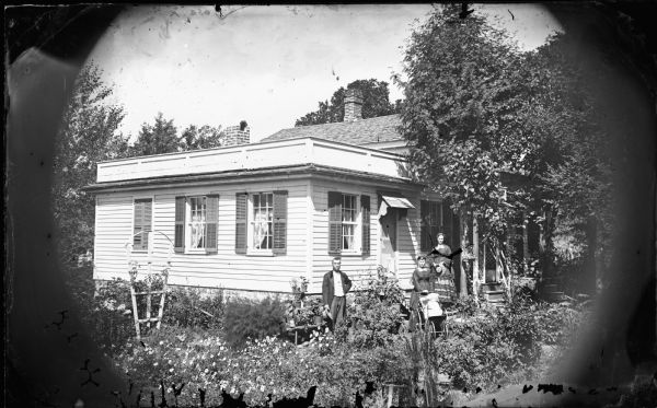 A family is posed in their garden with a baby in its buggy and a woman holding her hat. The frame house behind them has a parapet roof, a porch with latticework siding, and a screen door on the side.