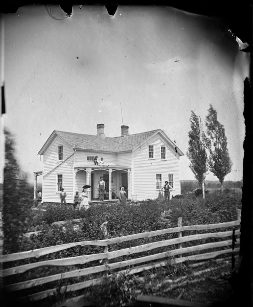 In the foreground is a fence, behind it a family posed in a yard. A child and dog are on the porch roof of the frame house behind the family.