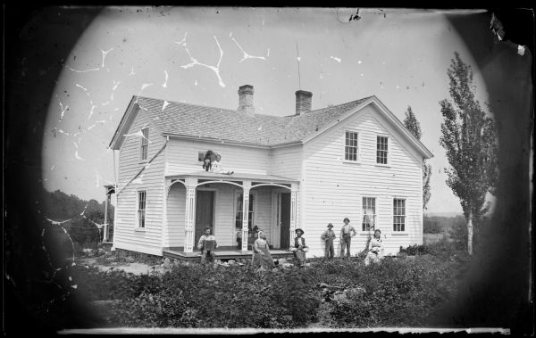 A family is posed in front of a wood frame house. A boy or girl with a doll is sitting with a dog on the porch roof. A man holding an accordion is sitting on the left side of the porch.