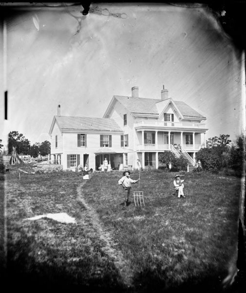 Allen Adsit stands in the left foreground with a pitch fork and his father, Stephen Adsit, is seated to the right. On the left near the house are two grindstones and a well. Women are posing near the porch on the left, and on the steps to the second-story porch on the right.
