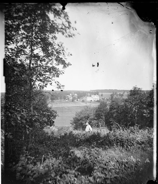 Allen E. Adsit is standing on a grassy hillside in the foreground.  In the distance is the Allen E. Adsit farm, which includes a three-story frame house with a porch and several farm buildings.