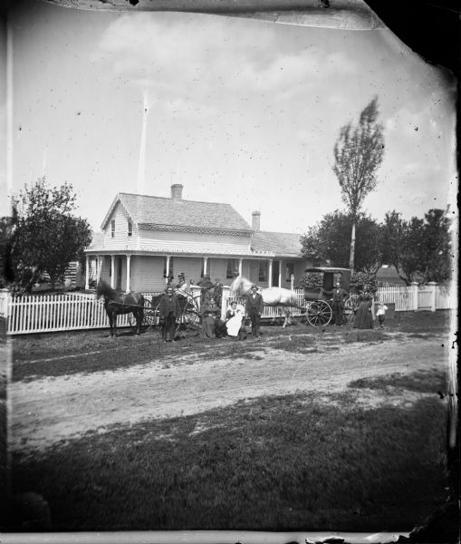 Family and frame house of Lars David Reque. Two carriages and a family are posed in front of a picket fence. Behind them is a large frame house with carpenter's lace trim. A wooden barrel is in the background.