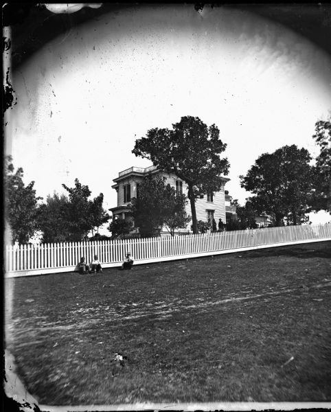 Three boys sitting in front of a picket fence. Behind them are more people in the yard of an urban bracket style house that has a small front porch, parapet roof and closed shutters.