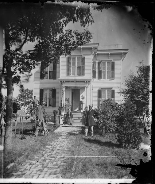 Two men, one holding a sword, and a woman are posing on the steps of a two-story bracket style house with a parapet roof and brick foundation. In the background at the left end of the house is a man holding a broad-brimmed cowboy hat.