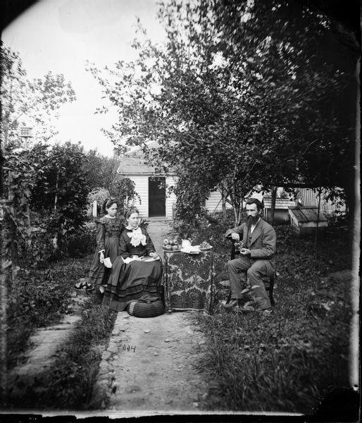 Benjamin F. Hahn, his wife Mary and their servant Sarah Hill sit around a table with an intricately patterned cloth with bowls of apples and grapes. The woman rests her feet on a hassock and the man holds a bunch of grapes. Behind them are a dog house, and a birdhouse on a post. Their frame house is obscured by trees. The farm is located in section 19 NE 1/4.