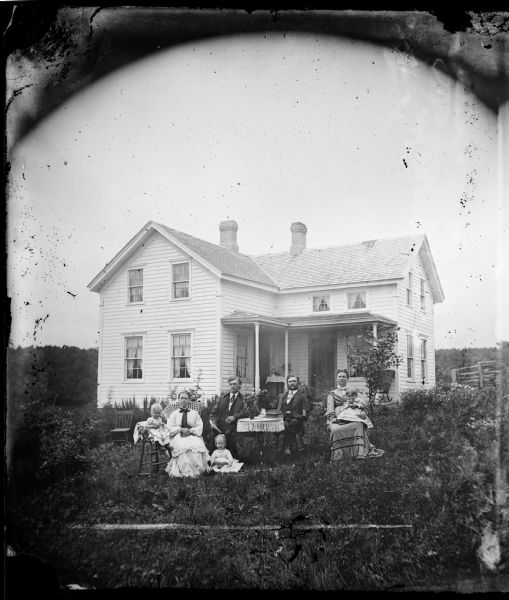 The P. Gunhouse family is posed around a table in their yard. The baby sits in a high chair and the table is covered with books and flowers. The L-shaped frame house behind them has a rocking chair on its porch and half windows just under the porch roof. A split-rail fence is at the far right.
