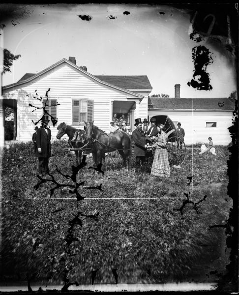 The Reverend Peter Andreas Rasmussen (1829-1898) is shaking the hand of a woman, possibly his wife Ragnhild Holland Rasmussen. Someone has apparently just arrived or is about to leave in a carriage. A boy sits behind them on a toy dog. Family members are posed on the porch of a one story frame house with a porch and small windows in back.