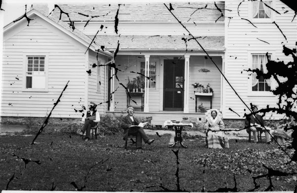 This is identified as the Jacob Larsen farm, but pictured are Ole Ashburn and his second wife seated in the yard with a table between them. The frame house behind them has a porch with two tables on it with plants in pots and two hanging plants. Clinton is located in the Jefferson Prairie settled by Norweigan immigrants.