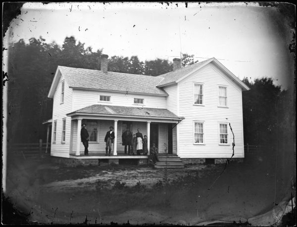 A family stands on the porch of an upright and wing frame house that has small second story front windows and a stone foundation.