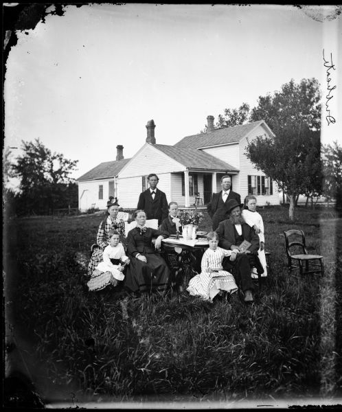 A family dressed in their Sunday clothes are gathered around a table that has flowers on it. The frame house behind them has shutters.