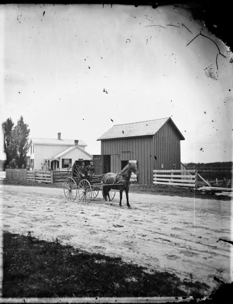 A couple sits in a carriage on a road adjacent to fenced-in farm buildings and an upright and wing frame house.