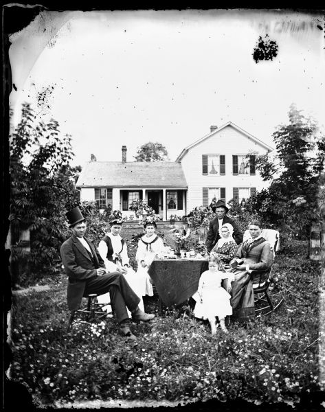 A family dressed in their finest clothes sits around a table with glassware on it. The man on the left wears a top hat at a jaunty angle. Behind them is a frame house with a porch on which are potted plants.