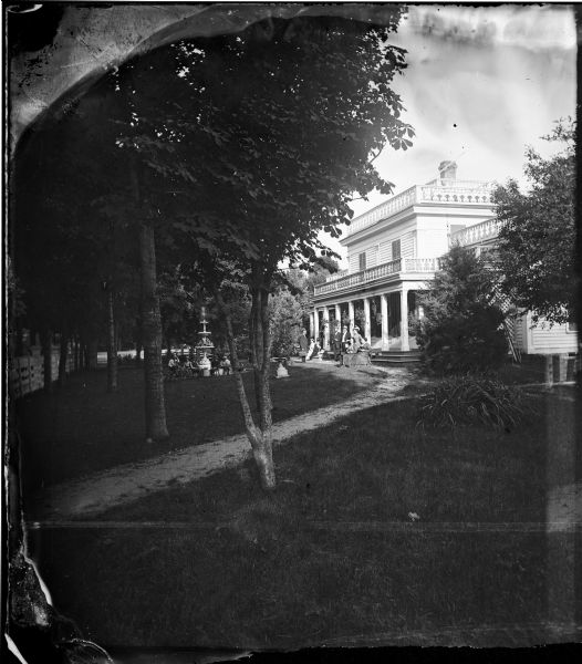 Family around fountain in fenced yard, with a boy on tricycle and family on steps of large frame house with parapet trim on porch and house roof. There are hanging plants on the porch and latticework on the porch side.