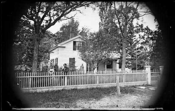 A family stands behind a picket fence in the yard of a frame Greek Revival house with latticework on its porch. There are two wood trellises in the yard.