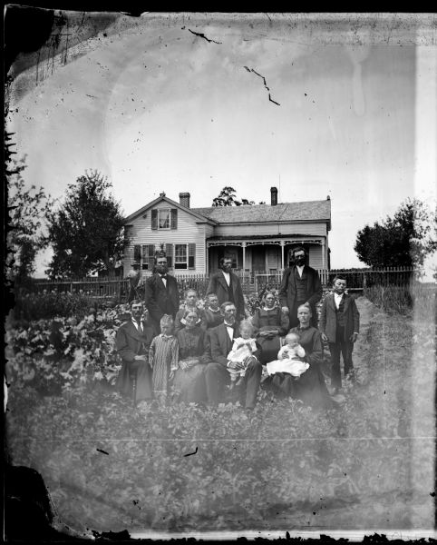 A large family group is posed in the foreground in front of a frame house with shutters and a lightening rod. Greek Revival style house, possibly belonging to Andres Maldstad. Behind them a boy hoes a garden.