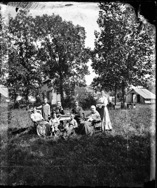 A family poses in a yard around a table. The grandmother appears to have an embroidery hoop in her lap. In the background on the right is a grindstone. Further back is a frame house with shutters, and farm buildings in the background on the right. "Williumson 12" is written on the negative, probably a misspelling of the name Williamson.