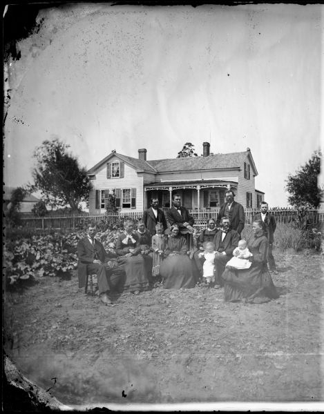 Fourteen members of what may be the Andres Malsted family pose in the garden. Behind them is a picket fence and behind it their upright and wing frame house that has some Greek Revival features.