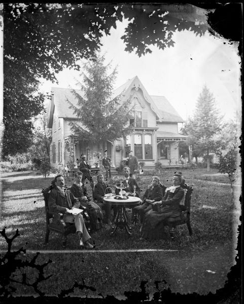 A very formal family is seated around a table ornamented with glassware. Men strike poses in the yard. Behind them is a large wood frame Victorian house with a bay window and wood trim, some of it in a Gothic Revival style. There is a double porch with hanging plants.