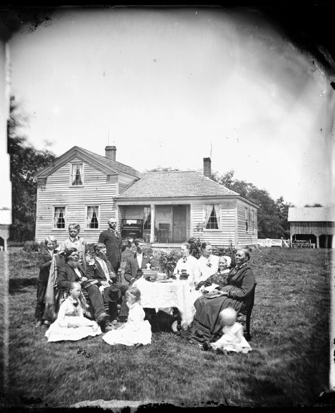 A family sits in a yard around a table with a girl holding an opened hand fan. The wood frame house behind them has a piano on its porch.