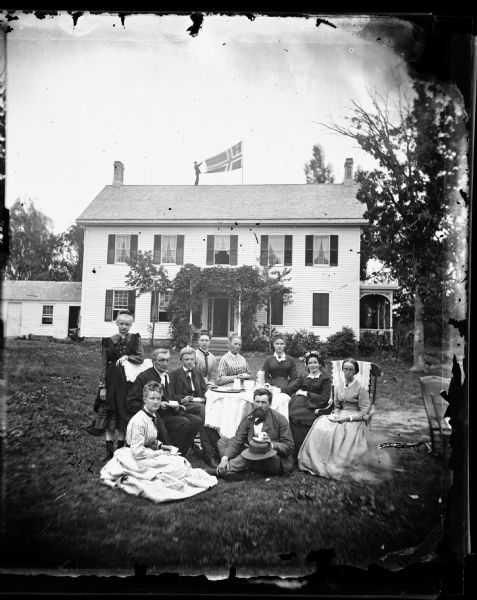The Reverend Jakob Aall Ottesen (1825-1904), his wife Cathinka Doderlein Ottesen and family posed in front of their home. A man on the roof holds out a Norwegian flag.