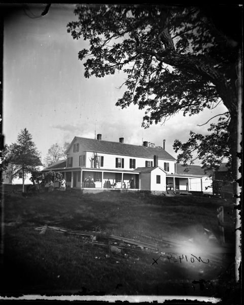 Ladies sit on the front porch of a long two-story house. One woman is next to a baby carriage. On the porch's roof is a man with a ladder painting the house. More people are in the yard including a man on a horse. A dinner bell sits atop a tall pole at the right end of the house.