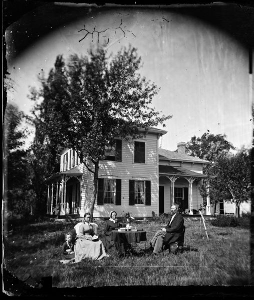 A man, two women, and a girl are posed in a yard by a table. The frame house behind them has cut work trim on its side and front porch and brackets on open spaced posts on both porches. The people are likely to be Jerome Skaalen, his wife Rebecca and a daughter.